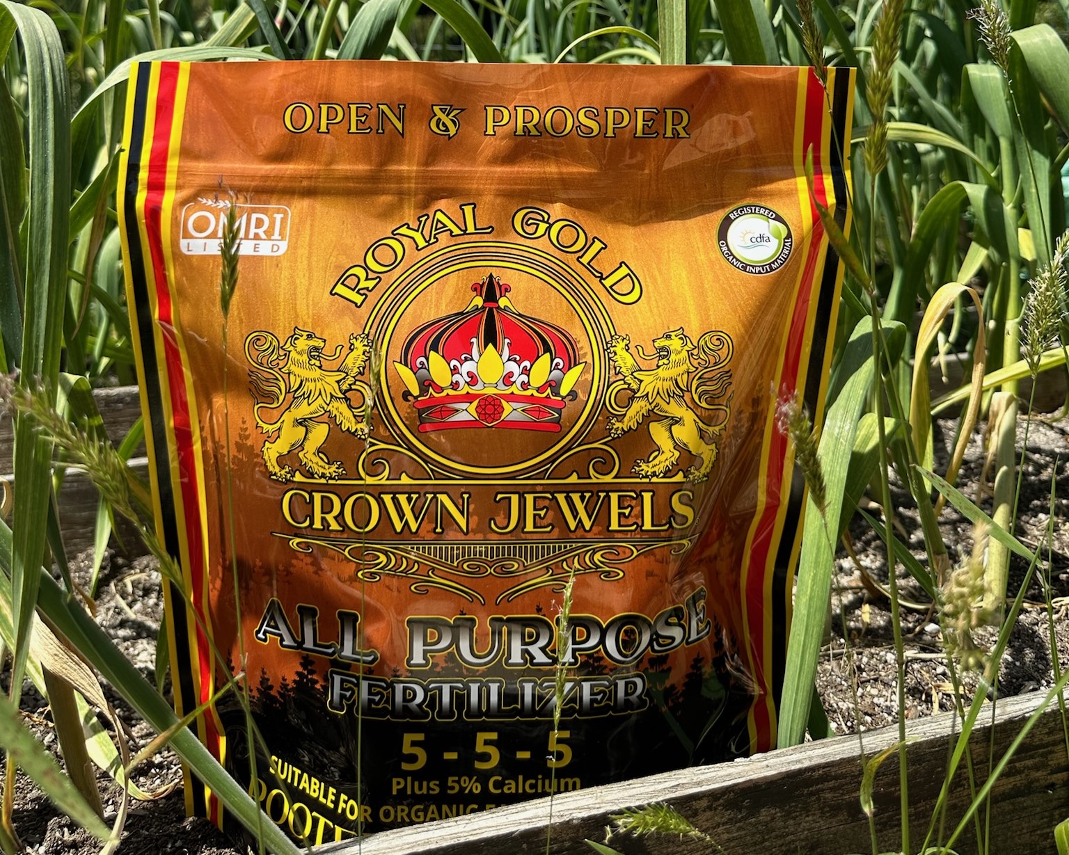 A bag of Royal Gold Crown Jewels All-Purpose 5-5-5 Fertilizer in a garden bed of garlic.